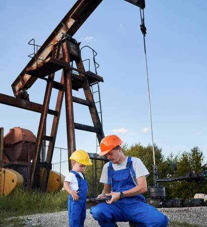 Engineer father introducing his son to work tool. Adult man and preschool child in workwears and helmets together looking to adjustable wrench on background of pump for oil production.